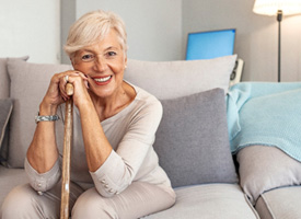 Woman in beige clothes on beige couch leaning on cane smiling